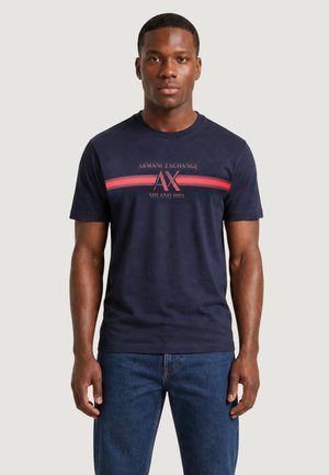 Mężczyzna ubrany w granatowy t-shirt Armani Exchange z czerwonymi paskami i napisem, w połączeniu z niebieskimi dżinsami, stoi na jednolitym jasnym tle.