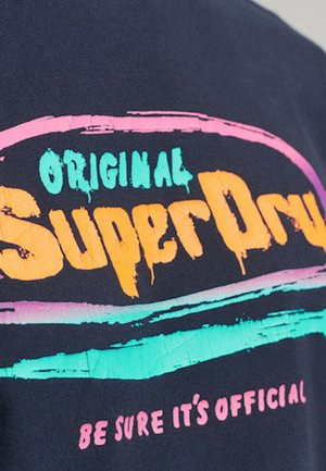 Ciemnoniebieska koszula z kolorowym napisem "Original SuperDry" w kolorze pomarańczowym, turkusowym i różowym oraz mniejszym różowym napisem "Be sure it's official" poniżej.
