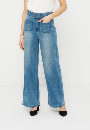 Donna che indossa jeans a vita alta blu a gamba larga con grandi tasche frontali, una cintura e tacchi neri a punta, in piedi contro uno sfondo bianco.