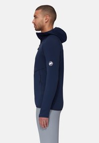 Giacca con cappuccio blu navy, realizzata in tessuto liscio, presenta una tasca con zip, un logo sulla manica e un design aderente.