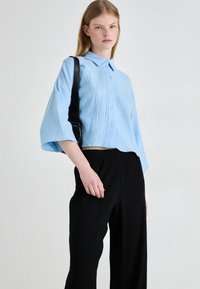 Camicetta azzurro chiaro, texturizzata, con colletto e chiusura a bottoni, abbinata a pantaloni neri larghi. Borsa a spalla nera indossata.