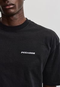 Voksen iført en sort t-shirt med hvid "PEGADOR" tekst på brystet mod en ensfarvet lys grå baggrund.