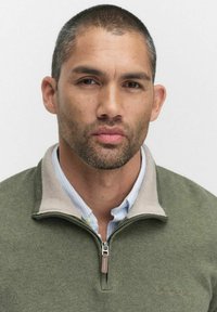 Grön fleece zip-up tröja med en beige krage. Modellen bär en ljusblå randig skjorta under. Enkelt design, inga mönster eller detaljer.