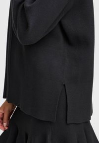 Schwarzer Pullover mit weichem Material, lässiger Passform und seitlichen Schlitzen, kombiniert mit einem schwarz-gemusterten Rock für einen geschichteten Look.