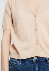 Hellrosa gestrickte Strickjacke mit kurzen Ärmeln, V-Ausschnitt und vier passenden Knöpfen; strukturierter, weicher Stoff. Kombiniert mit dunkelblauen Jeans.