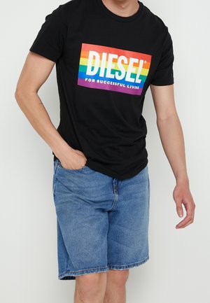Mężczyzna w czarnej koszulce z tęczowym logo "DIESEL for successful living" i niebieskich krótkich spodenkach jeansowych, stojący na jednolitym tle.