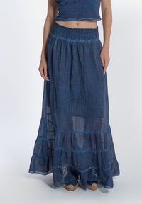 Gonna maxi in denim con un design a strati, vita smockata, texture morbida e orlo sfrangiato. Il colore è un blu profondo e ricco.