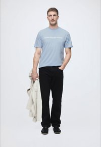 Ανοιχτό μπλε μπλουζί με κείμενο "DENIM DESIGN STUDIO", μαύρο παντελόνι, γκρι μπουφάν κρεμασμένο στο χέρι, μαύρα αθλητικά παπούτσια, ουδέτερο φόντο.