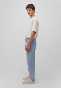 Hellgraues T-Shirt mit kurzen Ärmeln, lockere blaue Jeans und beige Sneakers mit roten Akzenten. Seitenansicht vor einem neutralen Hintergrund.