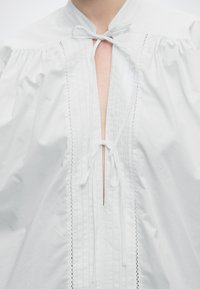 Blouse blanche avec manches bouffantes, plis verticaux et deux nœuds noués au niveau de l'encolure et de l'ouverture supérieure de la poitrine.