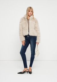 Kurtka z futerkiem w jasnym beżowym kolorze z zapięciem na zamek, zestawiona z ciemnoniebieskimi dżinsami typu skinny i czarnymi butami na obcasie typu slingback, trzymająca białą torebkę.