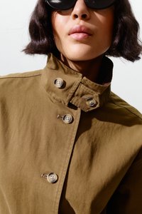 Naine, kes kannab ümmargusi musti päikeseprille ja pruunikat kõrge kraega nööbitud jopet, nähtaval on lühikesed tumedad lainelised juuksed.