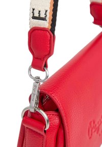 Punainen nahkainen käsilaukku, jossa on teksturoitu pinta, varustettuna kuvioidulla hihnalla ja hopeisella kiinnitysosalla. Brändin logo on painettu sivuun.