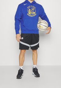 Felpa blu con grafica gialla, pantaloni corti neri con dettagli grigi e sneakers nere, mentre tiene un pallone da basket con dettagli bianchi e dorati.