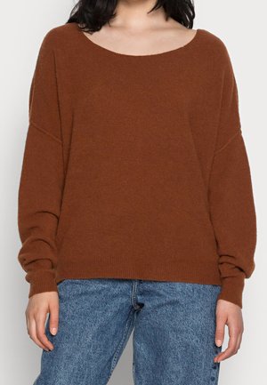 Femme portant un pull en maille brun ample à manches longues et un jean bleu en denim, debout devant un fond uni.