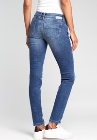 Jeans ajustados en denim azul medio, con un diseño clásico de cinco bolsillos, costuras visibles y un leve desvanecimiento. Combinados con zapatillas blancas.