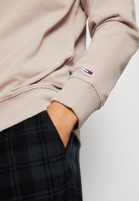 Beżowa bluza z prążkowanymi mankietami, z małym haftowanym logo w kolorach czerwonym, białym i niebieskim. W połączeniu z ciemnymi spodniami w kratę.