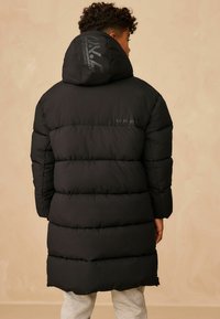 Čierny zateplený páperový kabát s kapucňou, s prešívaným designom, hladkým povrchom a logom na zadnej strane, s dĺžkou do stredu stehna.