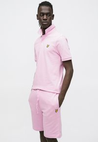 Rožinė polo marškinėliai su pakelta apykakle ir geltonu logotipu. Derinami su atitinkamais šortais su kišenėmis ir raišteliu. Lygios tekstūros.