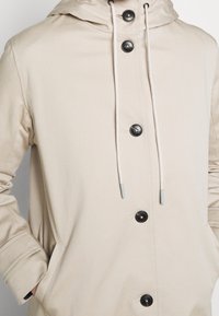 Beige jas met een capuchon met trekkoord, twee zijzakken en zwarte knopen. Gemaakt van een gladde stof met een minimalistisch ontwerp.