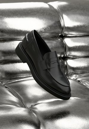 Mocasines de cuero negro con un acabado suave, una tira ancha en la parte superior y una suela elevada y texturizada. Presentados sobre un fondo metálico brillante.