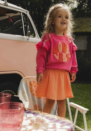 Felle roze sweatshirt met de tekst "GOOD VIBES", gerimpelde mouwen, gecombineerd met een gestreepte oranje rok. Buitenscène met een vintage voertuig.