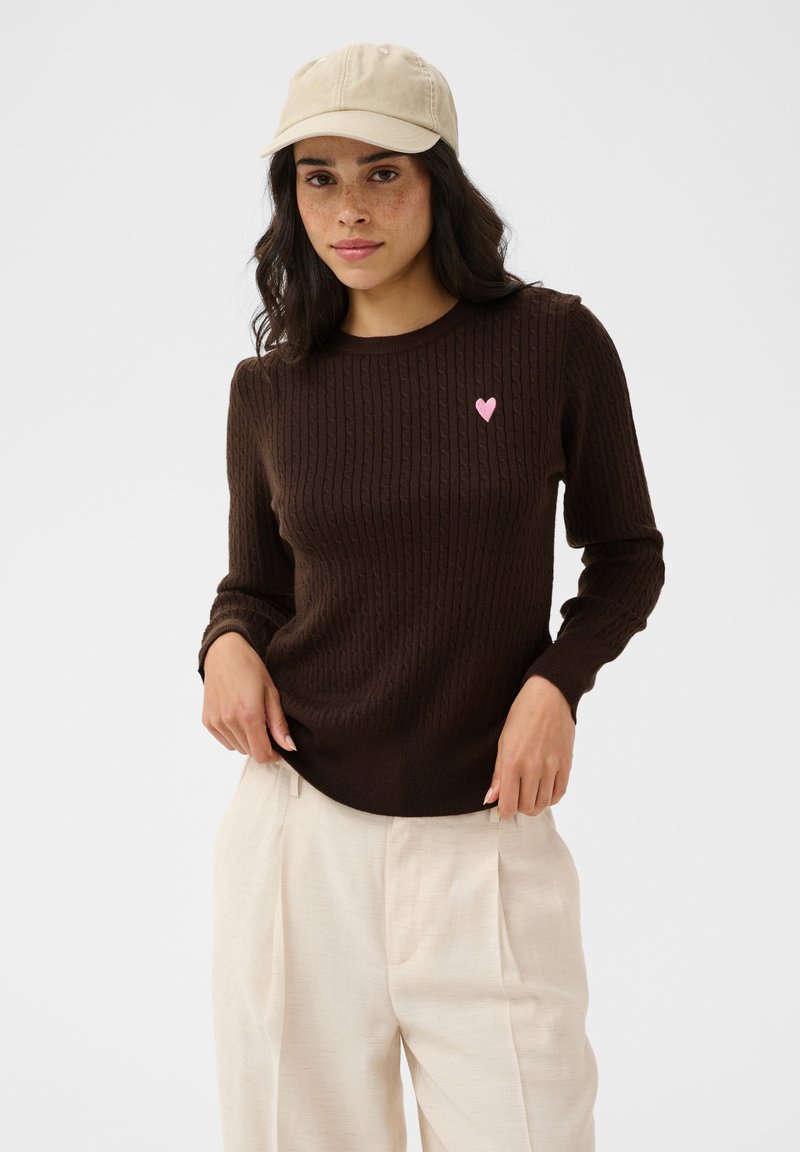 Femme portant une casquette beige, un pull torsadé brun foncé avec un petit cœur rose sur la poitrine et un pantalon plissé crème, debout devant un fond uni.