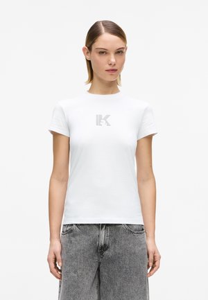 Femme aux cheveux raides porte un T-shirt blanc à manches courtes avec un logo "IK" gris clair et un jean noir décoloré taille haute, debout devant un fond uni.