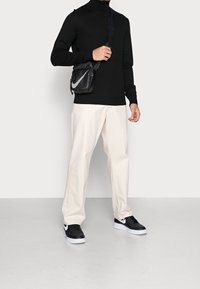 Zwarte gebreide coltrui, crèmekleurige broek met wijde pijpen, zwarte sneakers met witte zolen en een kleine zwarte schoudertas met een wit logo.