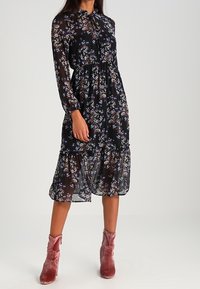 Mustaa kukkamekkoa, joka on valmistettu läpikuultavasta kankaasta, jossa on pitkät hihat, solmittava kaulus ja kerroksellinen helma. Yhdistetty vaaleanpunaisiin samettisiin nilkkabootseihin.