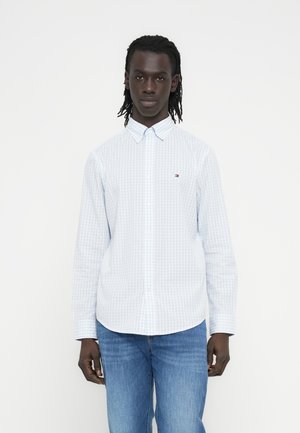 Junger schwarzer Mann mit Dreadlocks, der ein hellblau kariertes Hemd mit Knöpfen und blaue Jeans trägt, vor einem schlichten weißen Hintergrund stehend.