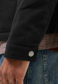 Manica di giacca in tela nera con chiusura a bottone a pressione. Dettagli di cucitura visibili e una camicia a quadri sotto. Mano con tatuaggi visibili.