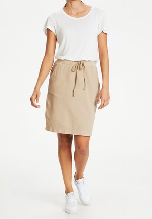 Femme marchant portant une chemise blanche à manches courtes, une jupe beige longueur genou avec lien à la taille, et des baskets blanches sur un fond blanc uni.