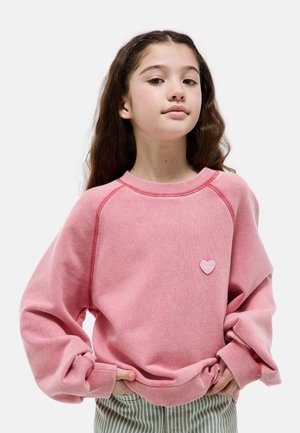 Fille aux longs cheveux bruns portant un sweat-shirt rose surdimensionné avec un petit cœur, les mains dans les poches avant.