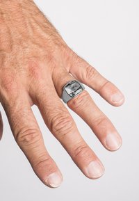 Ezüstring, négyzet alakú fekete márványbetéttel, egy enyhén szőrös bőrű kézen viselve. A körmök rövidek és természetesek.