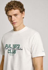 Balta medvilnė t-marškiniai su žaliais ir juodaisiais užrašais, įskaitant "PJL 1973", "Pepe Jeans London" ir "KLUBAS". Trumpi rankoviai, apvalios apykaklės dizainas.