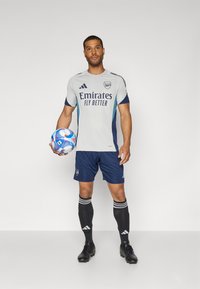 Grijze en marineblauwe voetbaloutfit met het logo "Emirates Fly Better", blauwe accenten, zwarte sokken met witte strepen, terwijl een kleurrijke voetbal wordt vastgehouden.