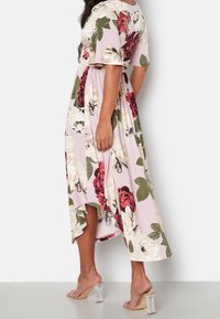 Femme portant une robe midi fleurie rose avec des fleurs blanches et rouges ainsi que des feuilles vertes, associée à des sandales à talons transparents sur fond blanc.