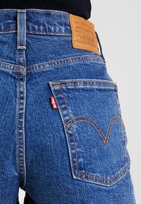 Blå denimjeans med hög midja, orange sömmar och en läderlogotyp. Har en distinkt design på bakfickan.