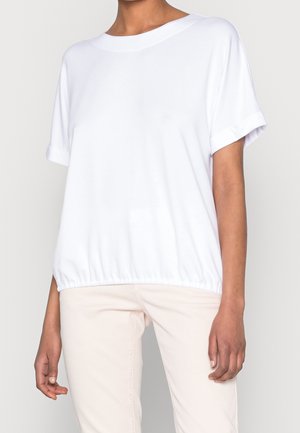 Personne portant un chemisier blanc ample à manches courtes avec ourlet élastique et un pantalon beige clair sur un fond uni.