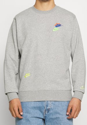 Uomo che indossa una felpa Nike grigio chiaro con numerosi piccoli loghi Nike in verde neon e arancione, abbinata a jeans blu.