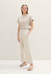 Top de manga corta beige combinado con pantalones de pierna ancha crema de cintura alta. Modelo con sandalias planas en tonos neutrales, de pie contra un fondo claro.