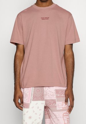 Mężczyzna w jasnoróżowej koszulce z napisem "Point Break Surf Club" oraz szortach w patchworkowy wzór w odcieniach różu i bieli.