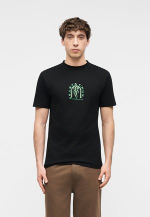 Młody mężczyzna z kręconymi włosami, ubrany w czarną koszulkę Santa Cruz Skateboards i brązowe spodnie, stoi na tle jednolitego jasnego tła.