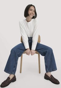 Witte blouse, blauwe wijde jeans en donkeren leren loafers. Zittend op een houten stoel met de handen op de knieën.