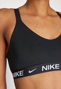 Crna sportska grudnjak s dubokim dekolteom, s podesivim trakama i debelim pojasom s natpisom "NIKE" u bijelim slovima. Glatka tekstura.