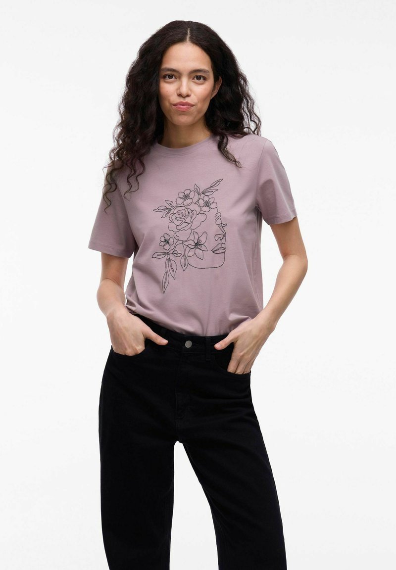 Lyserød bomuld t-shirt med en sort linjetegning af et ansigt omgivet af blomster. Sættet er parret med sorte bukser. Glat tekstur.