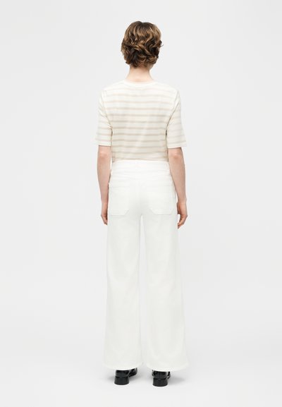Άτομο με κοντά κυματιστά μαλλιά που φοράει μπεζ ριγέ κοντομάνικο τοπ, λευκό παντελόνι ψηλόμεσο με φαρδιά πόδια και μαύρα παπούτσια, στραμμένο προς τα πίσω.