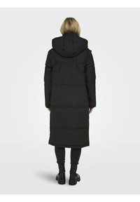 Musta pitkä puffer-takki hupulla, jossa on vuoratut osat ja vartalonmyötäinen siluetti, käytetty mustien legginsien ja jykevien saappaiden päällä.