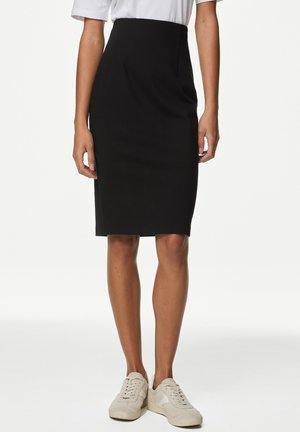Femme portant une jupe crayon noire jusqu'aux genoux, un haut blanc et des baskets beige clair, debout devant un fond uni.